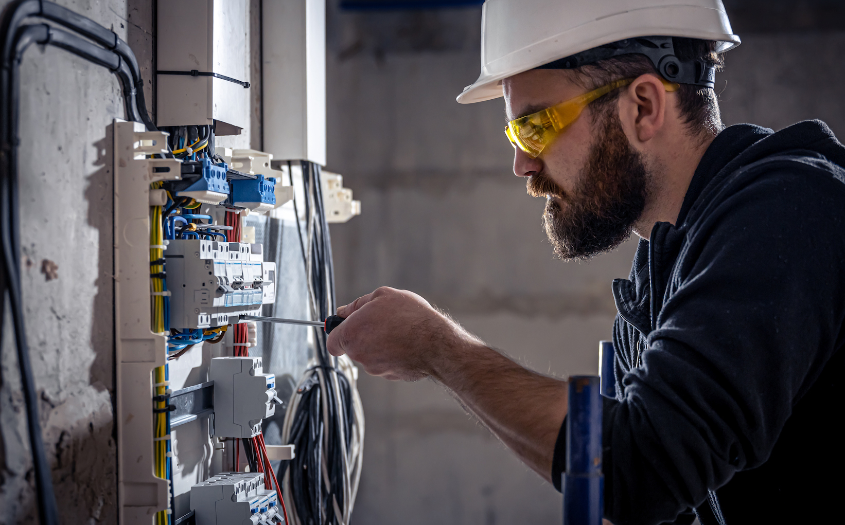 A male electrician works in a switchboard with an electrical connecting cable, connects the equipment with tools.